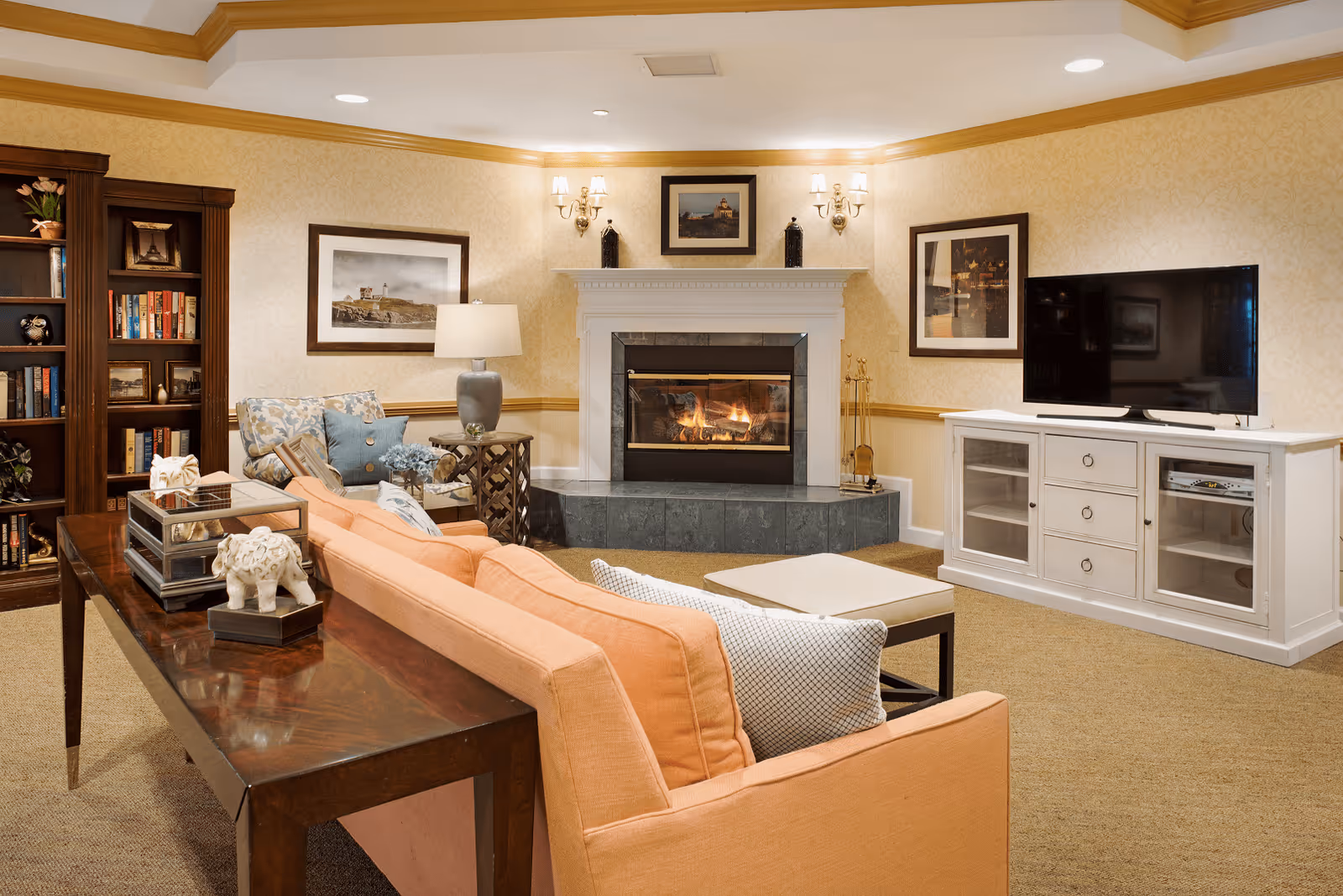 A cozy living room with a peach-colored sofa, a wooden console table with decorative items, a fireplace with a fire burning, framed pictures on the walls, a white TV stand with a flat-screen television, a bookshelf filled with books, and a comfortable armchair with a side table and lamp.