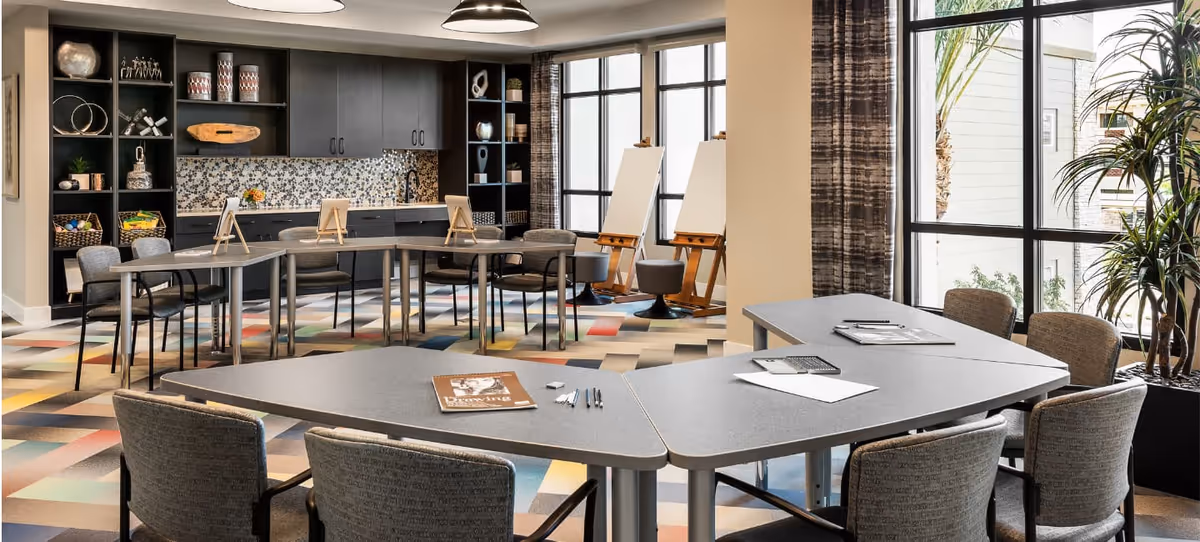 A bright and modern activity room with large windows letting in natural light. The room features several gray tables arranged in a U-shape with chairs around them. On the tables are magazines, pens, and paper. In the background, there are two easels with blank canvases, a colorful patterned carpet, and a wall with dark cabinets and open shelves displaying decorative items. There are also plants near the windows.