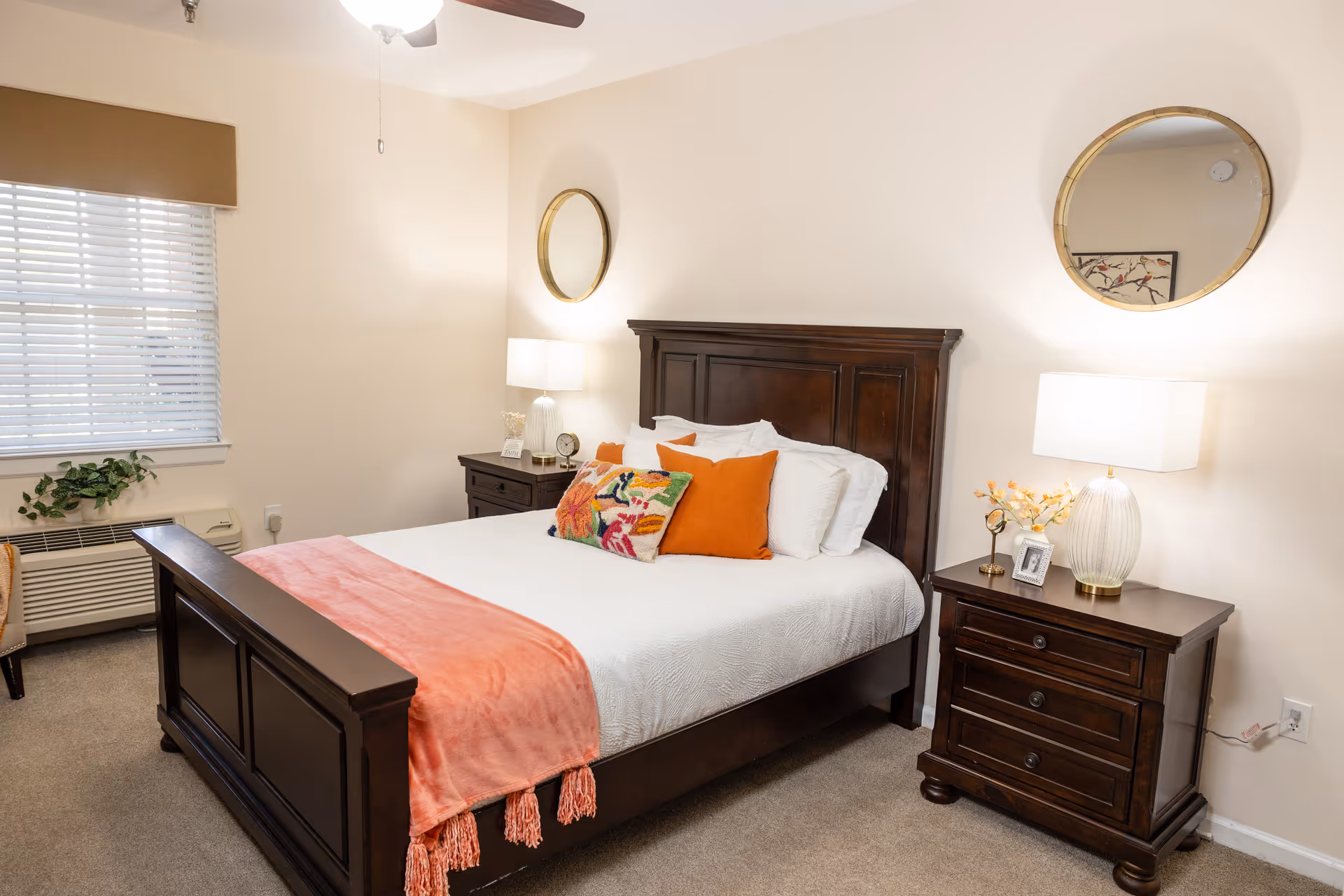 A neatly decorated bedroom with a dark wooden bed, white bedding and orange accents, flanked by matching nightstands with lamps and round mirrors on the wall.