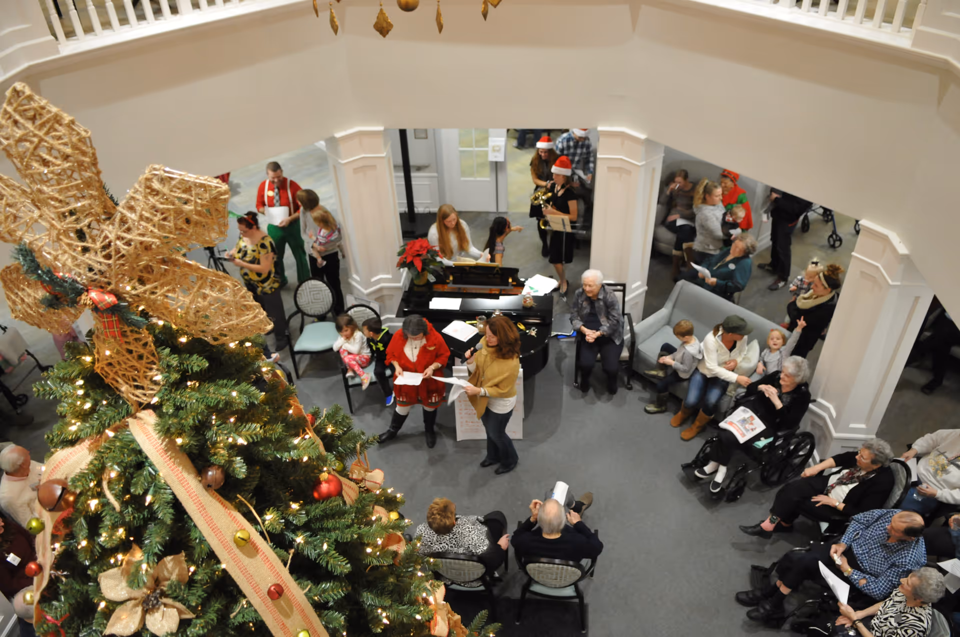 Overhead view of a festive gathering in a senior living facility with a large decorated Christmas tree in the foreground. Several elderly people and children are seated or standing around a piano where a woman is playing music. Some people are holding song sheets, and others are wearing holiday attire including Santa hats. The room has white walls and columns, and the atmosphere is lively and communal.