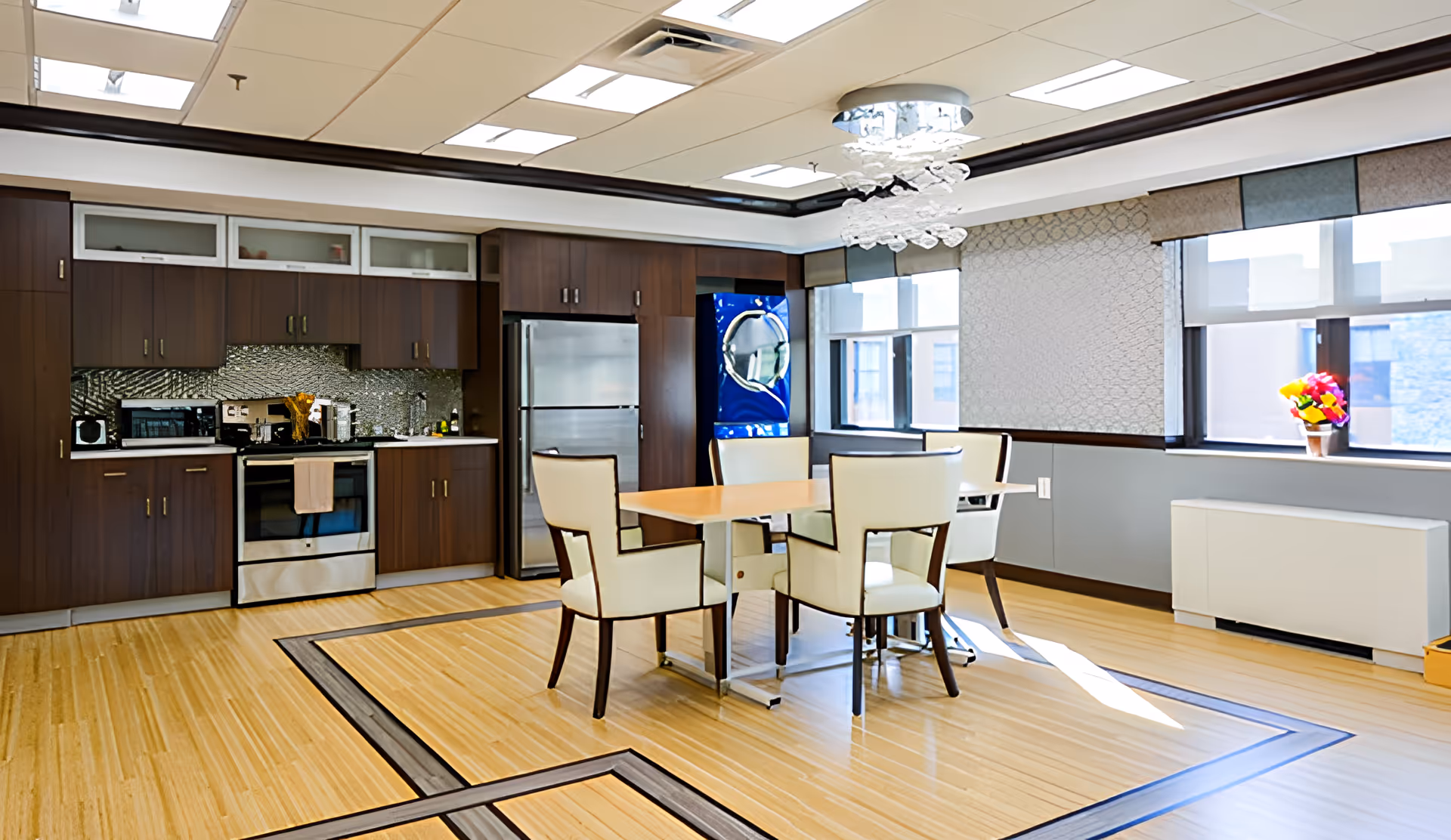 A modern kitchen and dining area with wooden cabinets, stainless steel appliances including a refrigerator and oven, a light wood dining table with four white cushioned chairs, large windows with patterned blinds, a decorative chandelier, and a small vase of colorful flowers on the windowsill.