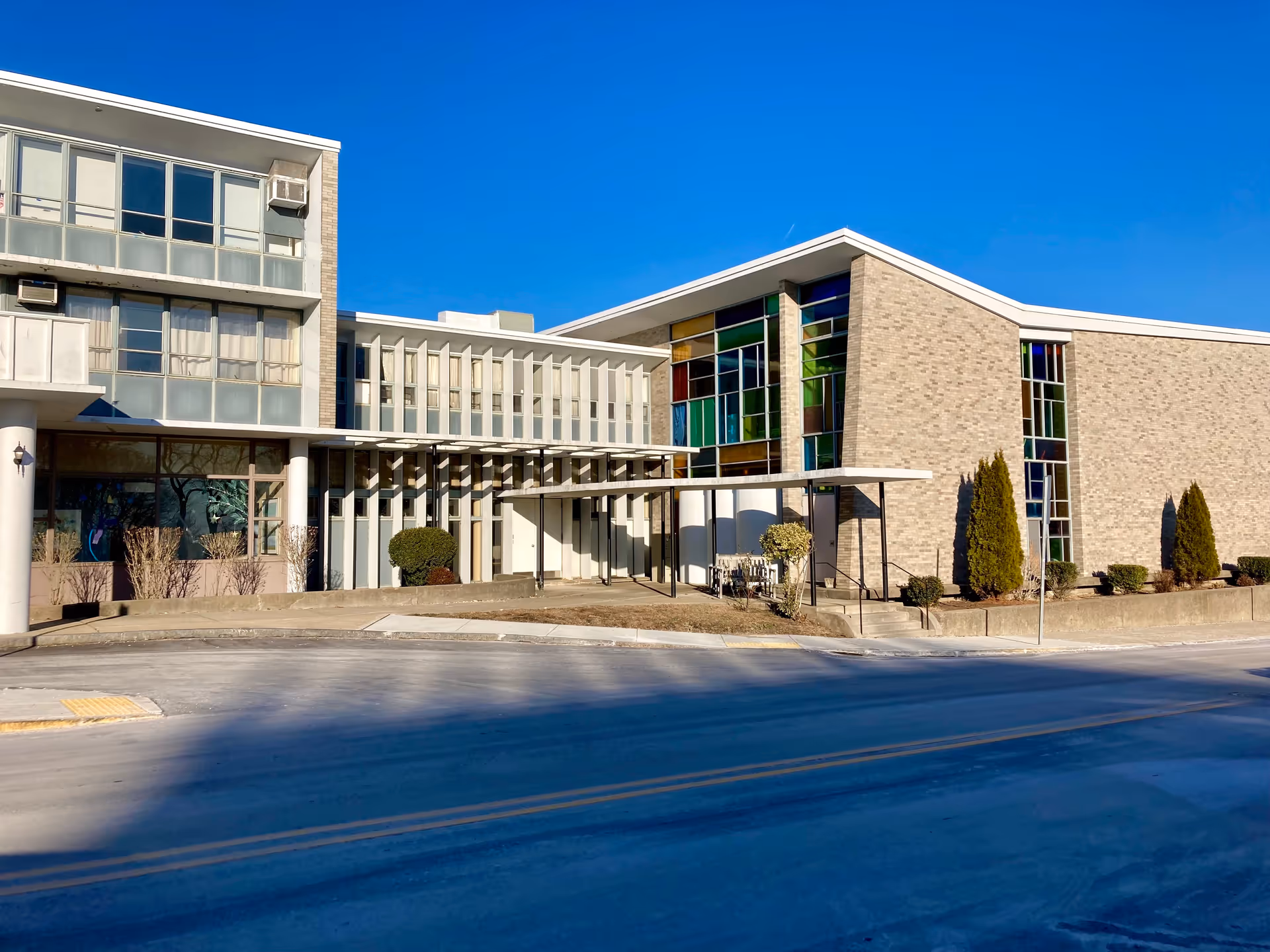 Exterior view of a modern building with large windows, some featuring colorful stained glass panels. The building has a covered entrance with a few small bushes and trees planted around it. The sky is clear and blue.