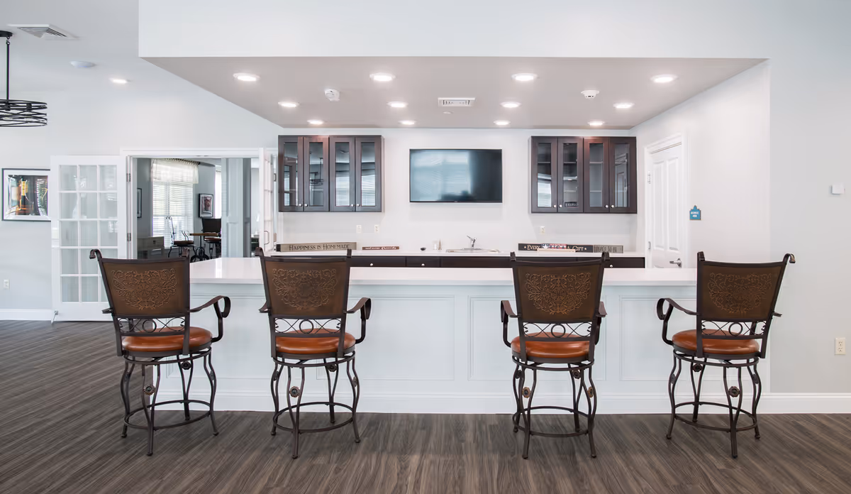 A modern kitchen area with a white island countertop and four high-back bar stools with brown leather seats and ornate metal backs. Behind the island are dark wood cabinets with glass doors, a mounted flat-screen TV, and a sink. The floor is a dark wood laminate, and the walls are painted light gray. To the left, there are glass double doors leading to another room with tables and chairs.