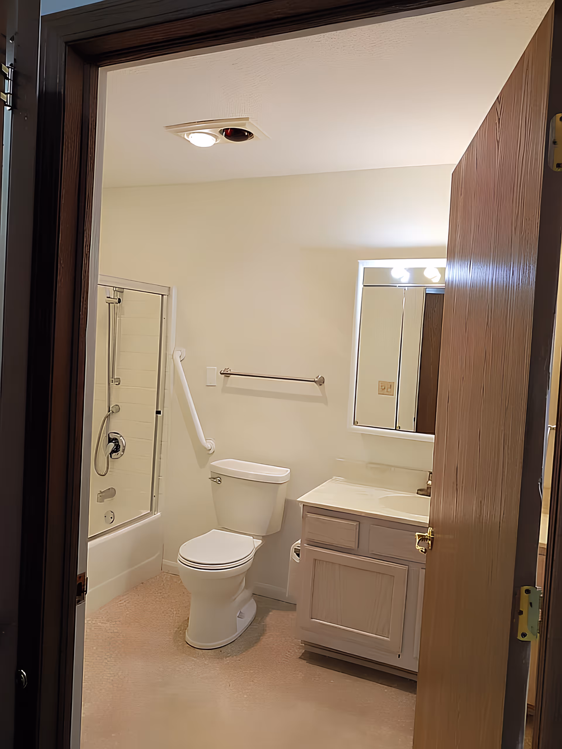View into a bathroom with a white toilet, a vanity with a sink and mirror above it, a towel rack, and a bathtub with a glass sliding door and a showerhead. The bathroom has light-colored walls and a wooden door partially open.