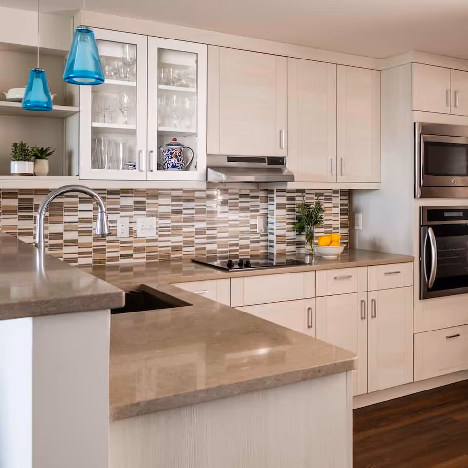 Modern kitchen with beige cabinets, a tiled backsplash in shades of brown and beige, a built-in oven and microwave, a stovetop with a stainless steel hood, a sink with a curved faucet, and two blue pendant lights hanging from the ceiling. There are glass-front cabinets displaying glassware and a bowl of fruit on the counter.
