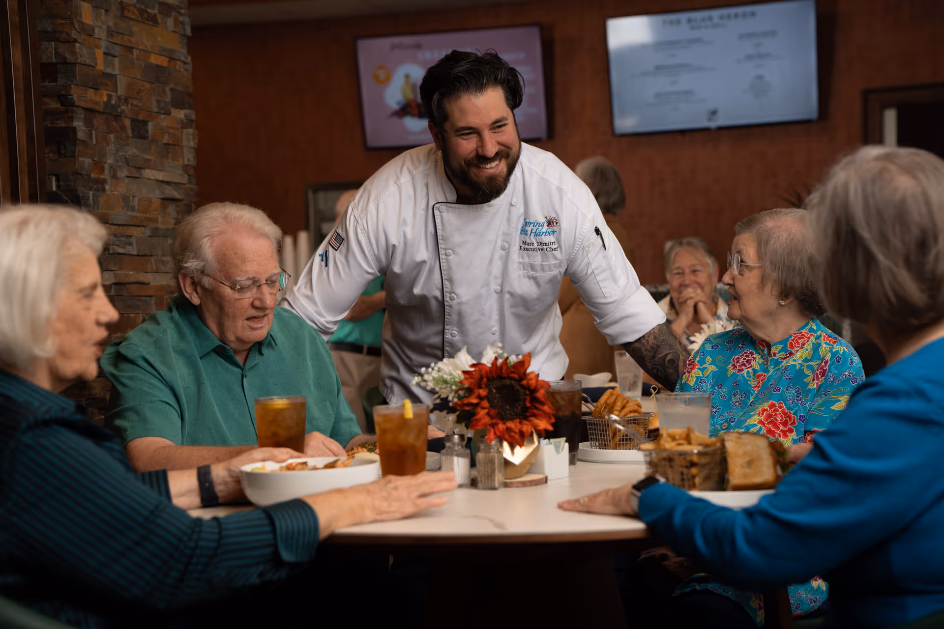 A smiling chef in a white uniform leans over a table and interacts with four elderly people seated around it, who are enjoying a meal and drinks in a cozy dining area with a stone wall and two TV screens in the background.