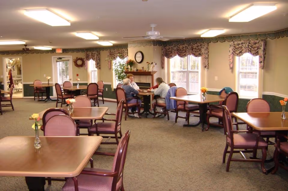 A dining room in Prairie Gardens Assisted Living with multiple tables and chairs arranged neatly. Each table has a small vase with flowers. There are large windows with floral valances allowing natural light to enter. In the background, a few elderly people are seated at a table near a fireplace with a clock above it. The room has carpeted floors and ceiling lights.