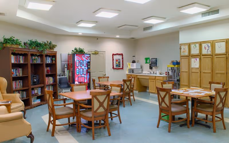 A well-lit common room with several wooden tables and chairs arranged for group seating. There is a bookshelf filled with books and plants on top, a vending machine, a microwave on a counter, and a popcorn machine. The walls have some children's artwork displayed, and the floor is covered with blue and beige tiles.