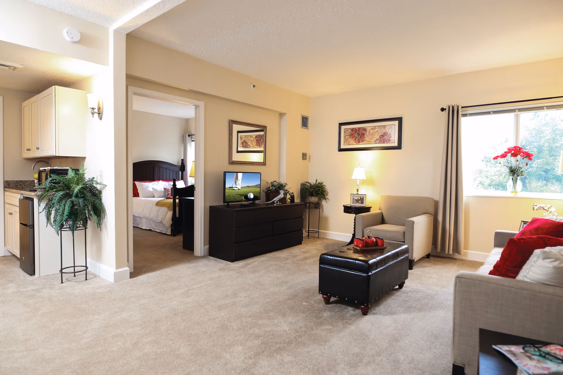 Bright, furnished living room of an assisted-living suite with a sofa, armchair, ottoman, TV dresser, window and a view into a bedroom and kitchenette.