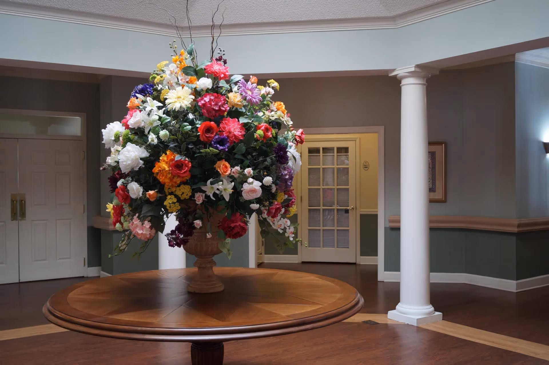 A large floral arrangement with various colorful flowers sits on a round wooden table in the center of a spacious room with hardwood floors, white columns, and muted green and beige walls. In the background, there are double doors and a glass-paneled door.
