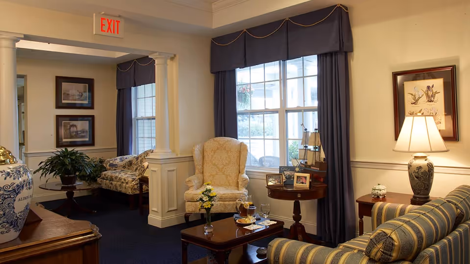 A cozy living room area with a striped sofa, a beige armchair, a wooden coffee table with a vase of flowers and drinks, a side table with framed photos and a model ship, a lamp on another side table, and large windows with dark curtains letting in natural light.