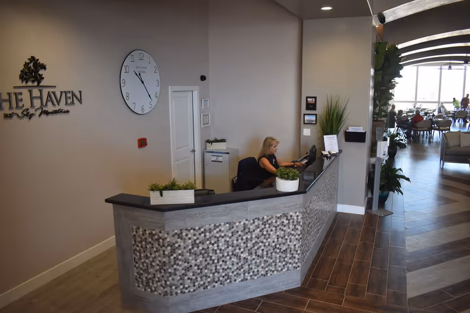 Reception area of The Haven at Sky Mountain Independent and Assisted Living Community with a woman working at the front desk. The desk has a decorative mosaic front and potted plants on top. A large clock is mounted on the wall behind the desk, along with the facility's name. In the background, there is a spacious common area with tables, chairs, and large windows letting in natural light.