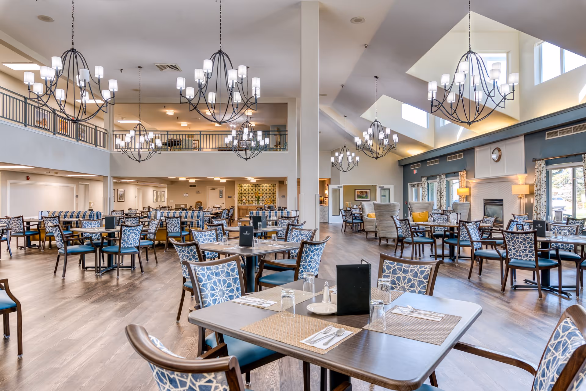 A spacious and well-lit dining room in a senior living facility with multiple tables and chairs arranged neatly. The room features high ceilings with large chandeliers, a fireplace, and large windows allowing natural light to enter. The decor includes patterned upholstery on chairs and a mix of seating options including armchairs and dining chairs.