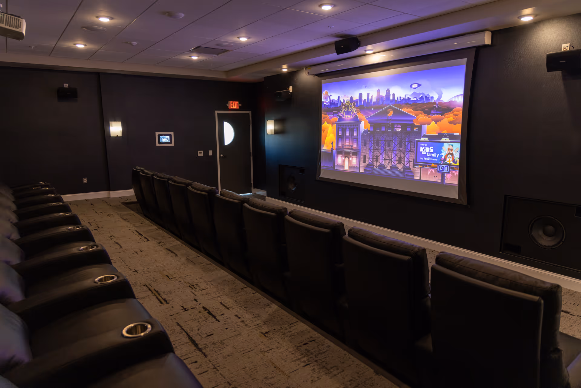 A small theater room with two rows of black leather recliner chairs facing a large screen displaying a colorful animated cityscape. The room has dark walls, recessed ceiling lights, wall sconces, and speakers mounted on the walls. There is an exit door at the back of the room.