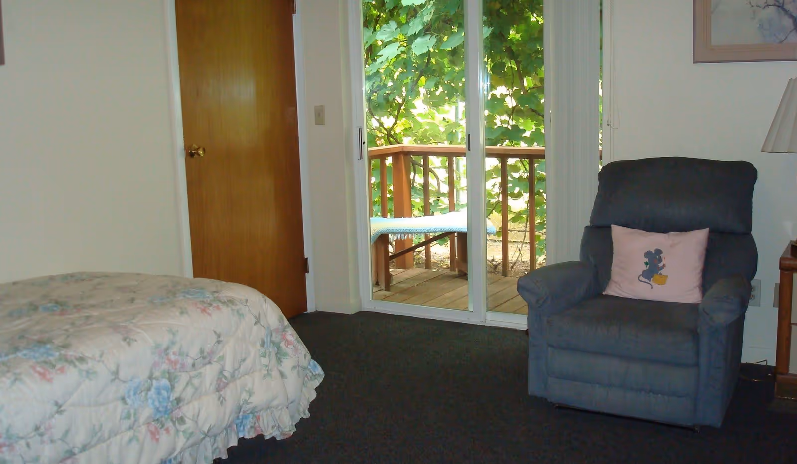 A cozy bedroom with a floral-patterned bedspread on the left, a wooden door in the background, and a sliding glass door leading to a small wooden balcony with greenery outside. On the right side, there is a dark blue recliner chair with a pink pillow featuring a cartoon mouse, and a wooden nightstand with a lamp.