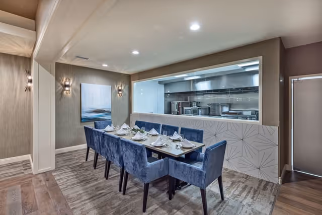Private dining room with a table set for eight, blue upholstered chairs, and a large window looking into a commercial kitchen.
