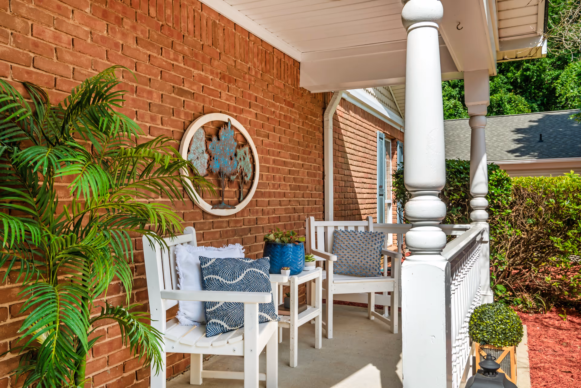 A front porch with white wooden chairs, patterned cushions, potted plants, and decorative wall art against a brick exterior.