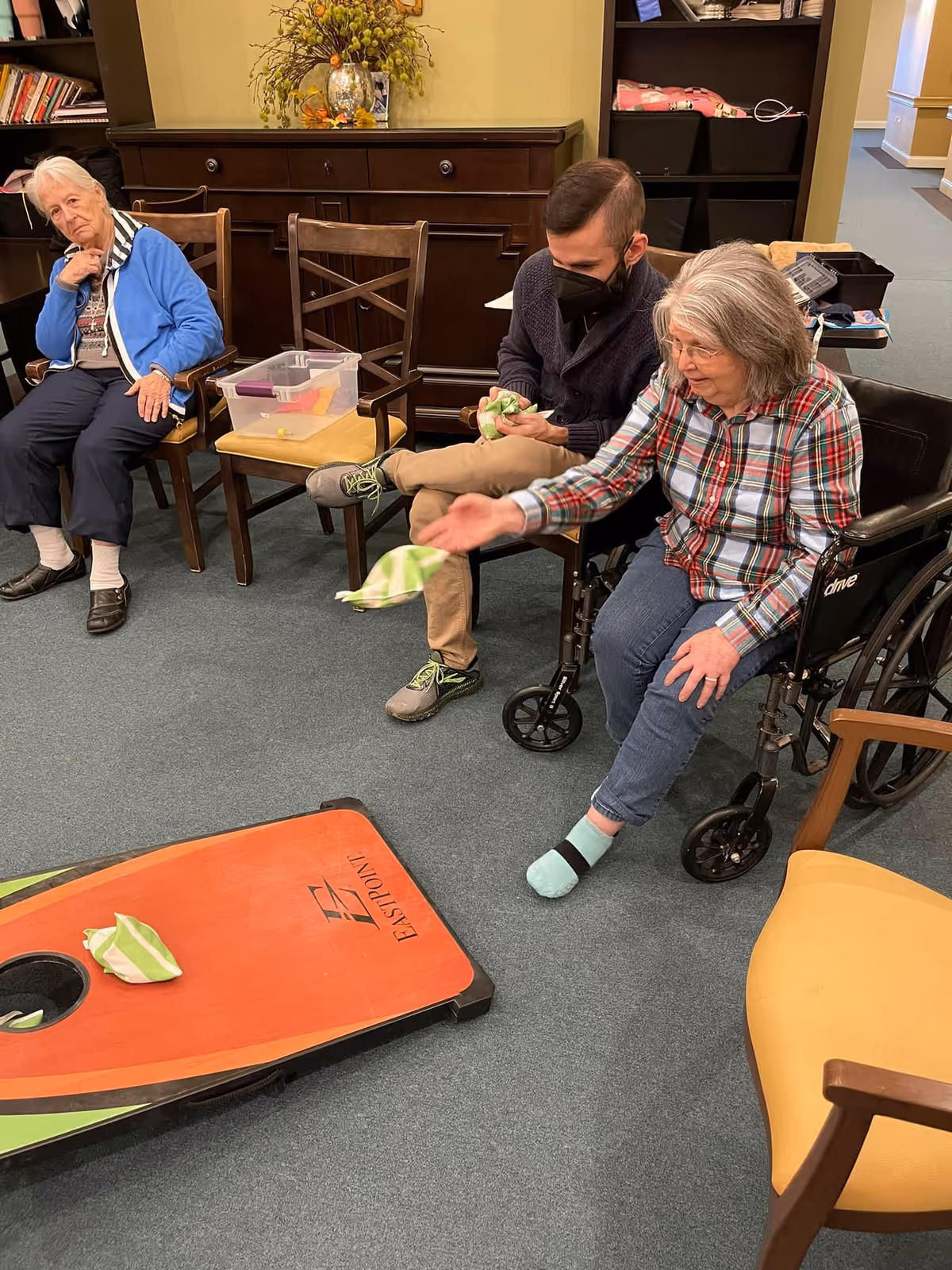 Three people in a room at San Gabriel Memory Care. An elderly woman in a wheelchair is tossing a bean bag towards a cornhole board on the floor. A man wearing a black face mask sits next to her, holding a bean bag. Another elderly woman sits on a chair nearby, watching the activity. The room has wooden furniture, including a cabinet and chairs, and a carpeted floor.