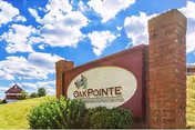 Brick-framed sign reading 'Oak Pointe Nursing & Rehab Center' set among shrubs with a blue sky and clouds overhead.