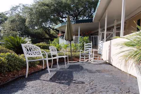 Sunlit outdoor patio with white wrought-iron chairs and umbrella beside a building.
