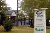 Outdoor view of The Senior Villages facility entrance with a sign displaying the facility name, contact number, and a gated area with trees and buildings in the background.
