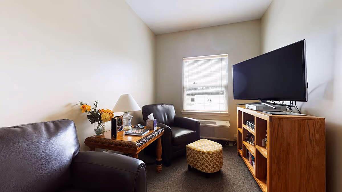 A small living room with two dark leather armchairs, a wooden side table with a lamp, flowers, and books, a patterned ottoman, a window with blinds, and a wooden TV stand holding a flat-screen television and books.