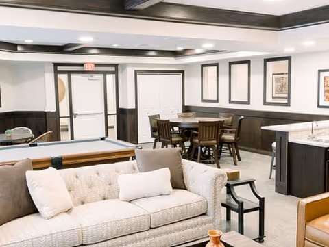 A well-lit common area in a senior living facility featuring a beige tufted sofa with white and brown pillows, a pool table, a round wooden table with chairs, framed artwork on the walls, and a small kitchen area with a countertop and bar stools.