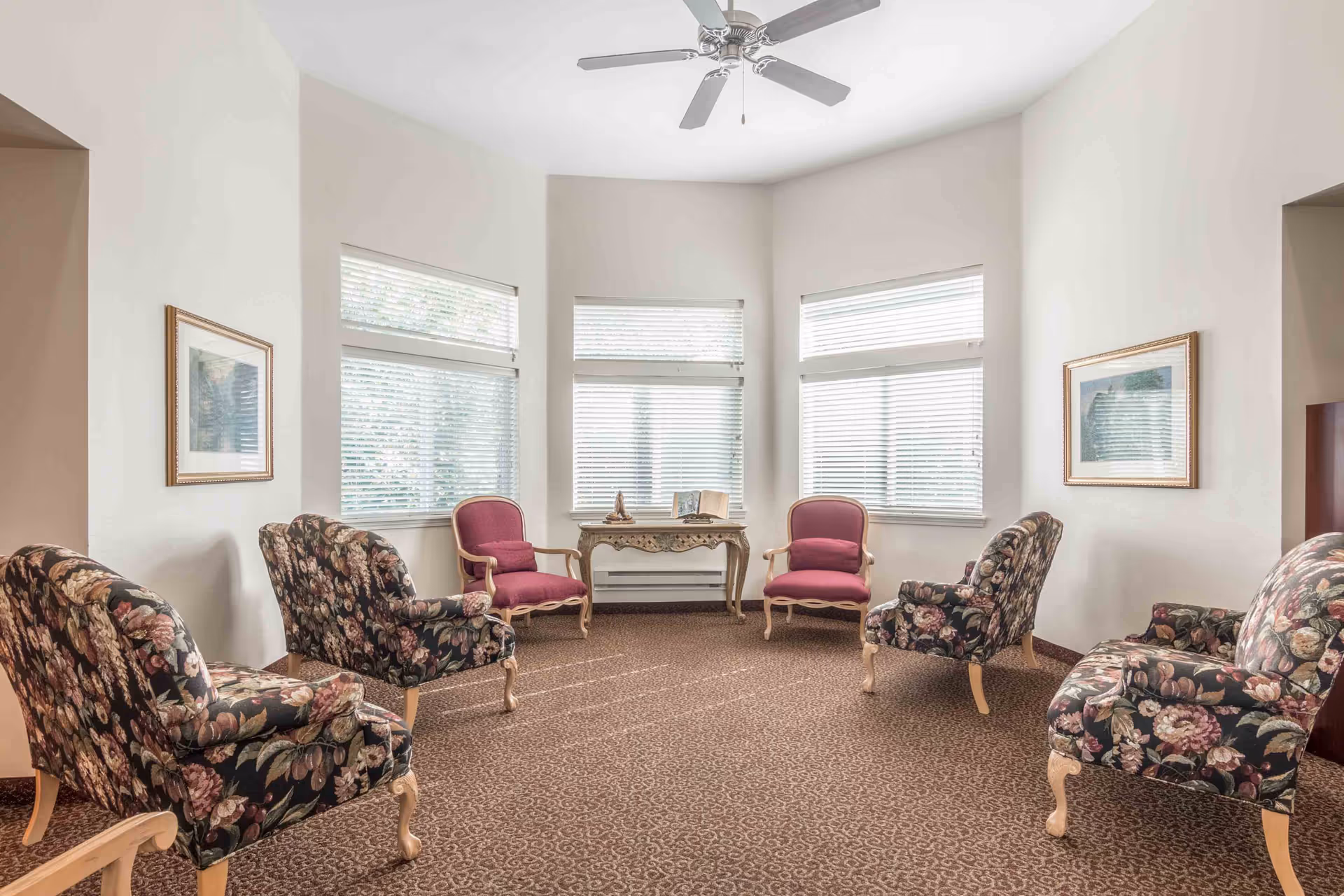 A bright sitting room with six upholstered chairs arranged in a circle on a patterned carpet. Four chairs have a floral pattern, and two chairs are solid pink with wooden frames. The room has three large windows with white blinds, two framed pictures on the walls, and a ceiling fan above. A decorative wooden table with an open book and a small sculpture is placed against the windows.