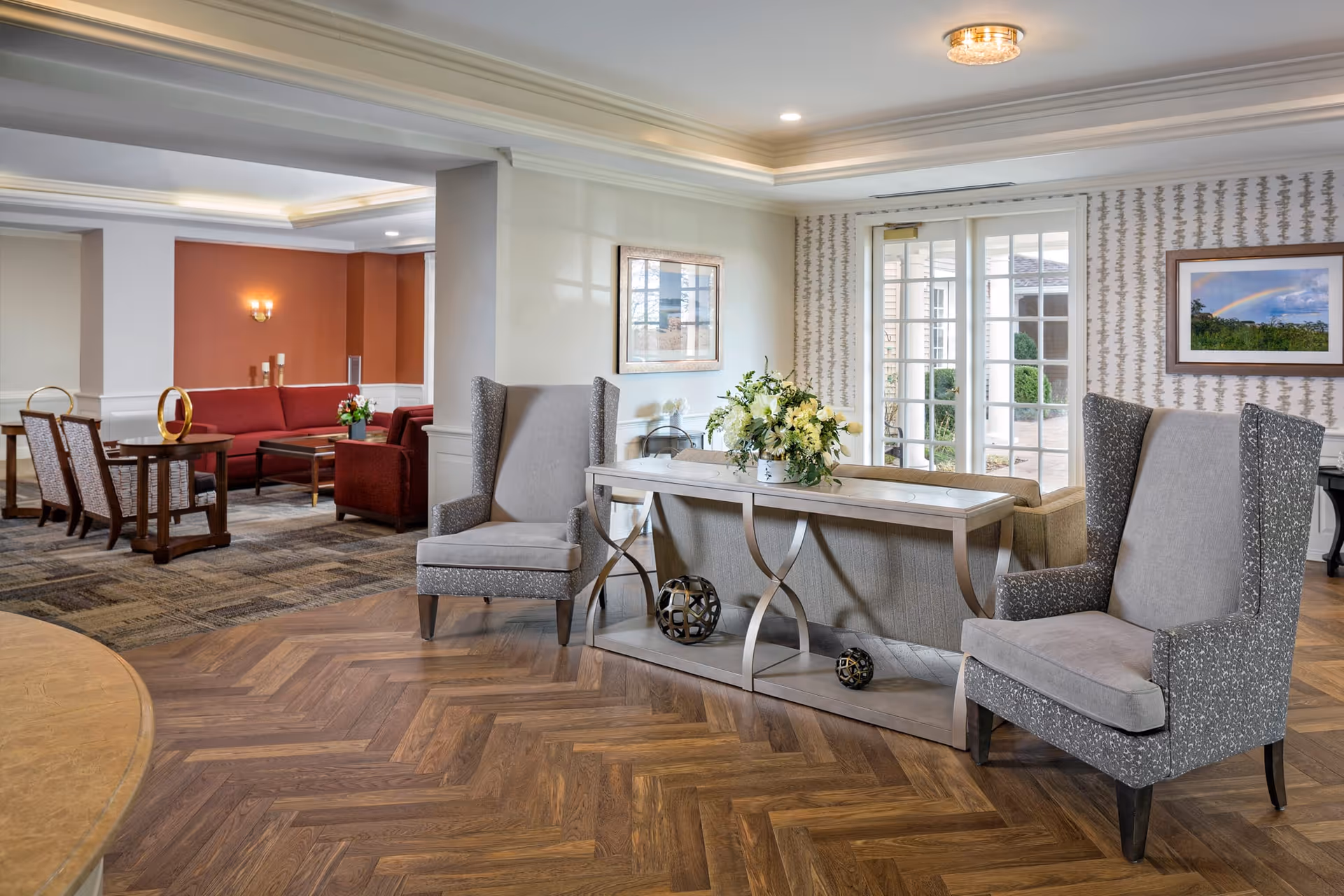 A spacious and well-lit living room area in a senior living facility with two gray upholstered armchairs facing a console table decorated with a flower arrangement and decorative spheres. In the background, there is a seating area with red sofas and wooden chairs, a patterned carpet, and framed artwork on the walls. Large glass doors lead to an outdoor patio area.