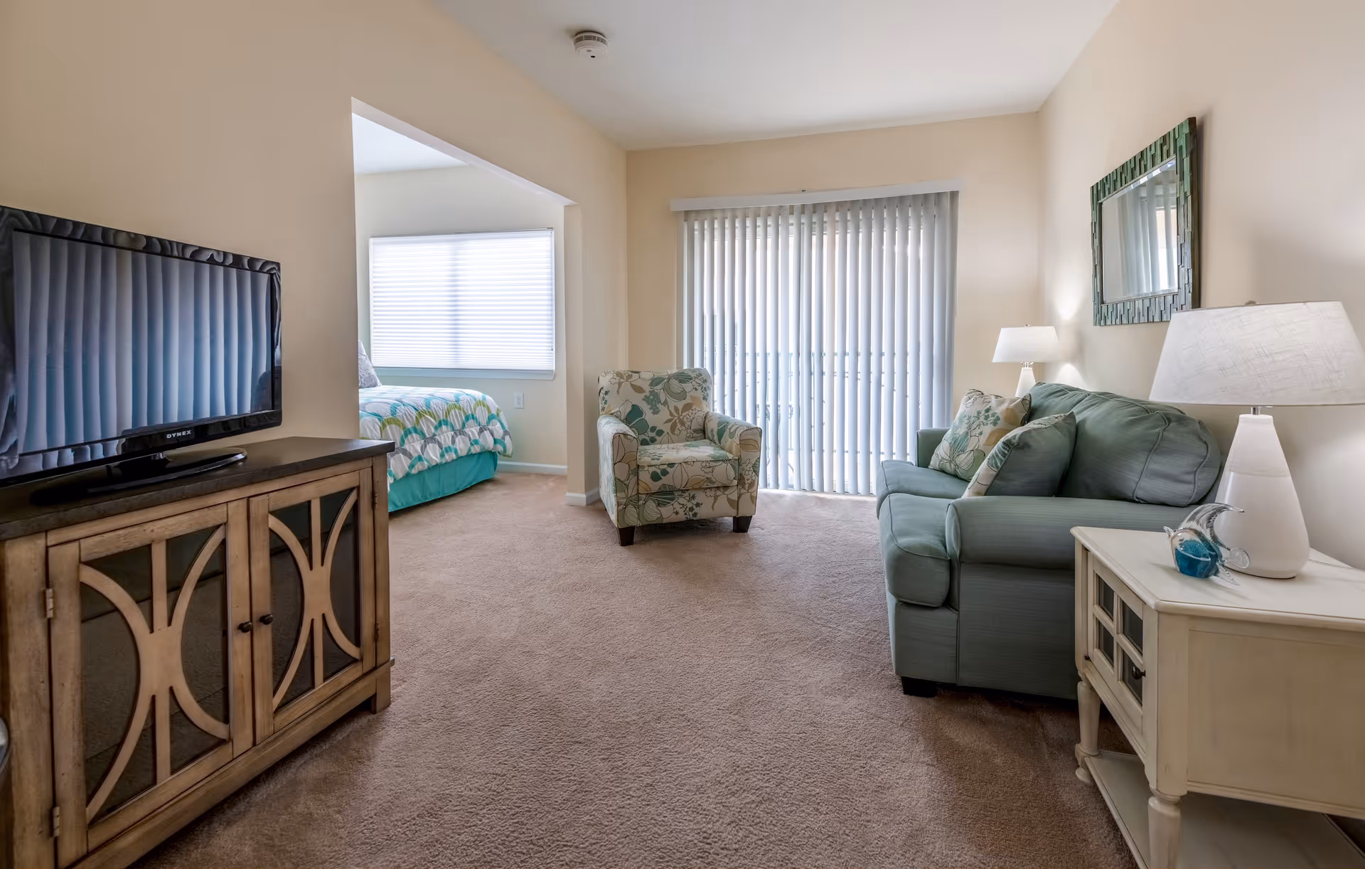A cozy living room area with a green sofa, a floral armchair, a white side table with a lamp, and a wooden TV stand with a flat-screen TV. The room has beige walls and carpet, with vertical blinds covering a sliding glass door. In the background, a bedroom with a bed and a window with blinds is visible.