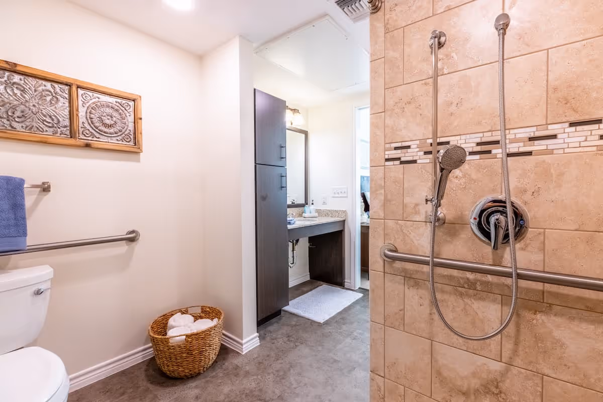 A clean and accessible bathroom featuring a white toilet with a blue towel hanging on a grab bar above it, a wicker basket with rolled white towels on the floor, and a tiled shower area with a handheld showerhead and grab bars. In the background, there is a vanity with a sink, a large mirror, and dark cabinetry.