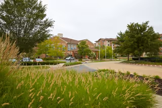 Front view of a senior living building with landscaped grasses, trees, and a parking area.