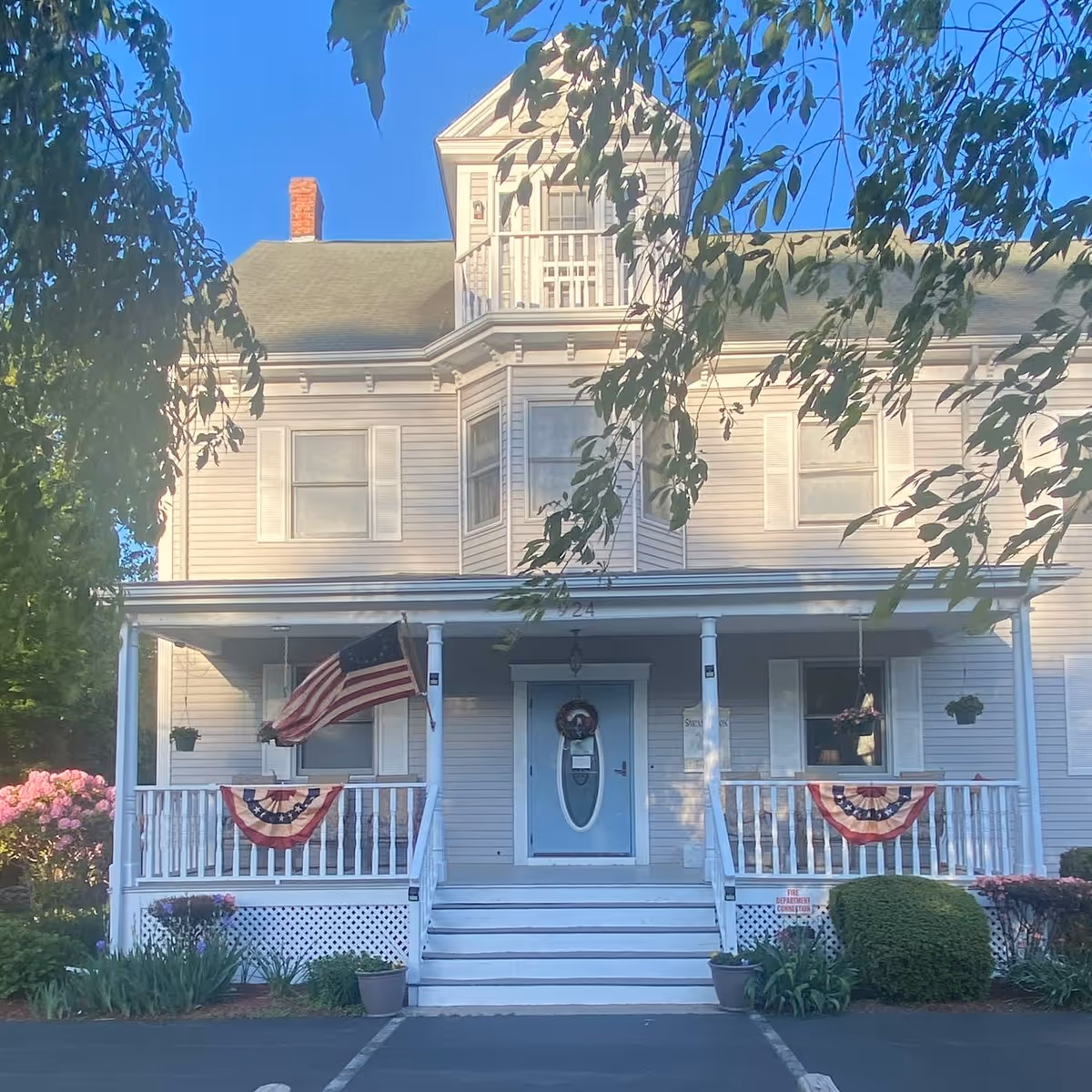 Front exterior view of a large, light gray, two-story house with a porch decorated with American flags and patriotic bunting. The house has white railings, a blue front door with an oval window, and a small balcony above the porch. There are plants and flowers around the porch and a clear blue sky in the background.