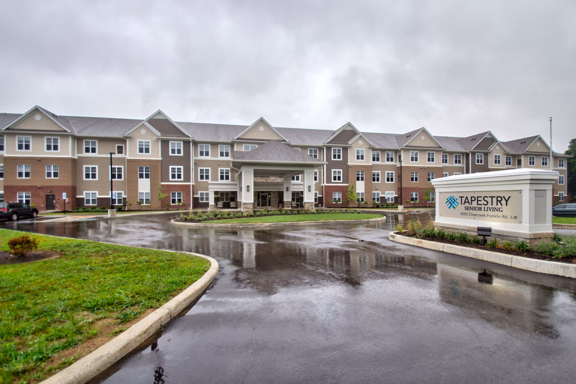 Exterior view of a large three-story senior living facility building with a covered entrance and a circular driveway. The building has multiple windows and a mix of beige, brown, and white colors. A sign in front reads 'Tapestry Senior Living' with the address 8683 Clearcreek Franklin Rd.