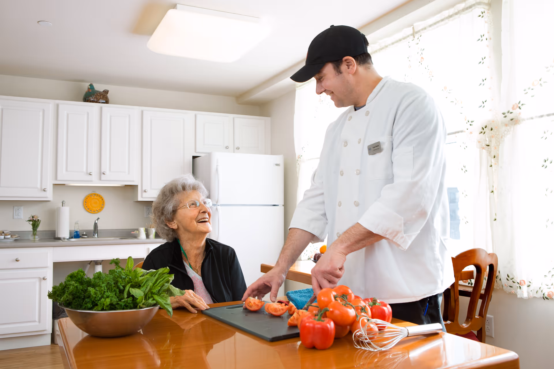 An elderly woman sitting at a kitchen table smiling and looking at a chef who is slicing tomatoes. The kitchen has white cabinets, a refrigerator, and a bowl of leafy greens on the table.
