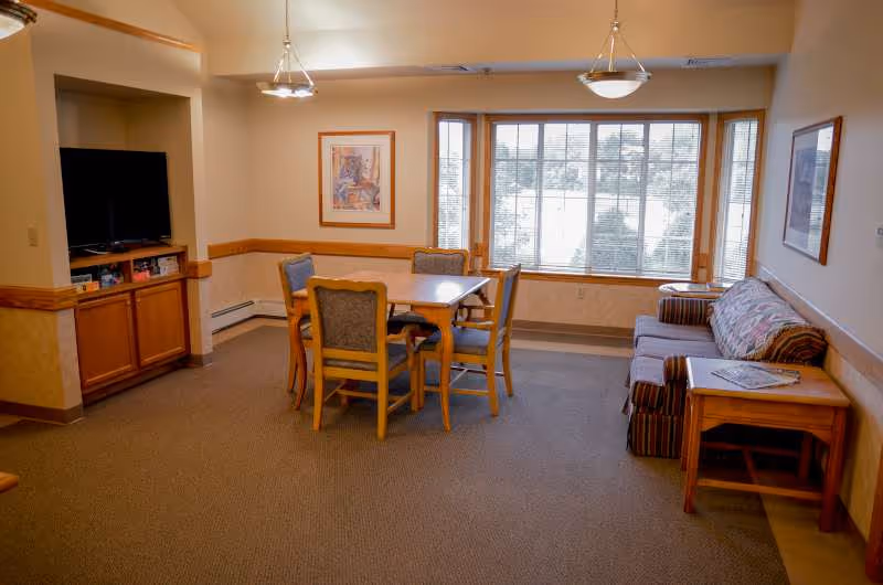 A cozy living room area with a wooden table and four chairs in the center, a couch with patterned cushions on the right, a side table with a magazine, a large window with blinds letting in natural light, framed artwork on the walls, and a TV set in a wooden cabinet on the left.