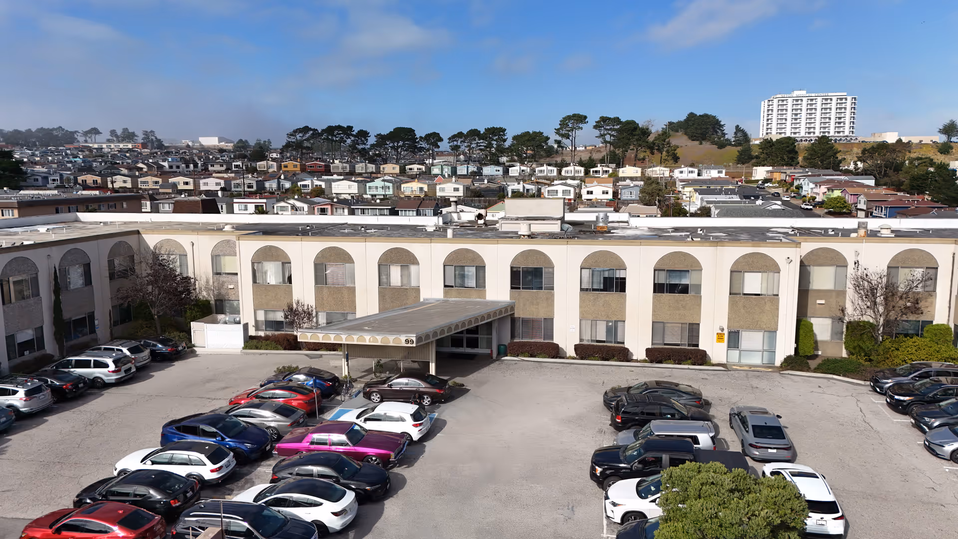 Exterior view of a long building with arched windows and a large parking lot in front.