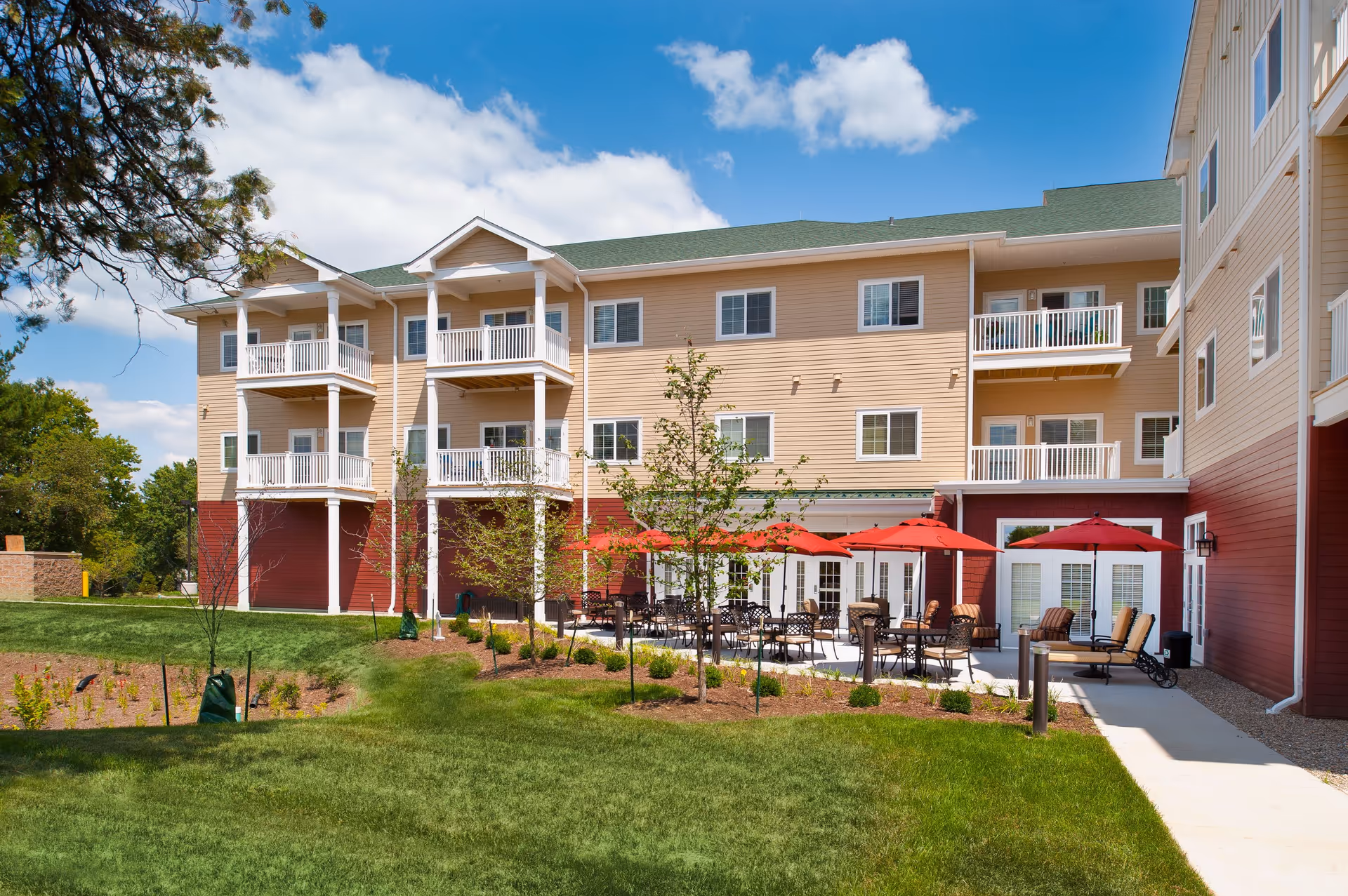 Outdoor patio area of a senior living facility with several tables and chairs under red umbrellas, surrounded by a well-maintained lawn and young trees. The building has beige and red siding with white balconies and a green roof under a partly cloudy blue sky.