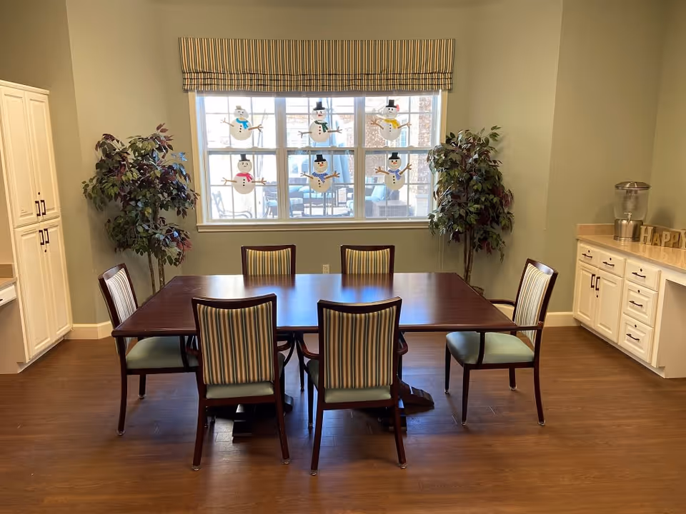 A dining room with a rectangular wooden table surrounded by six chairs with striped upholstery. Two artificial plants are placed on either side of a large window decorated with snowman cutouts. To the right, there is a countertop with cabinets underneath and a water dispenser on top.