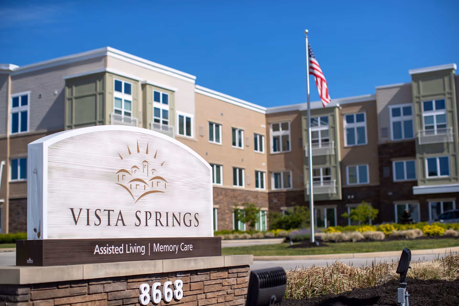 Exterior view of Vista Springs Greenbriar Village, an assisted living and memory care facility, showing the building facade, a large sign with the facility name and services, an American flag on a flagpole, and landscaping under a clear blue sky.