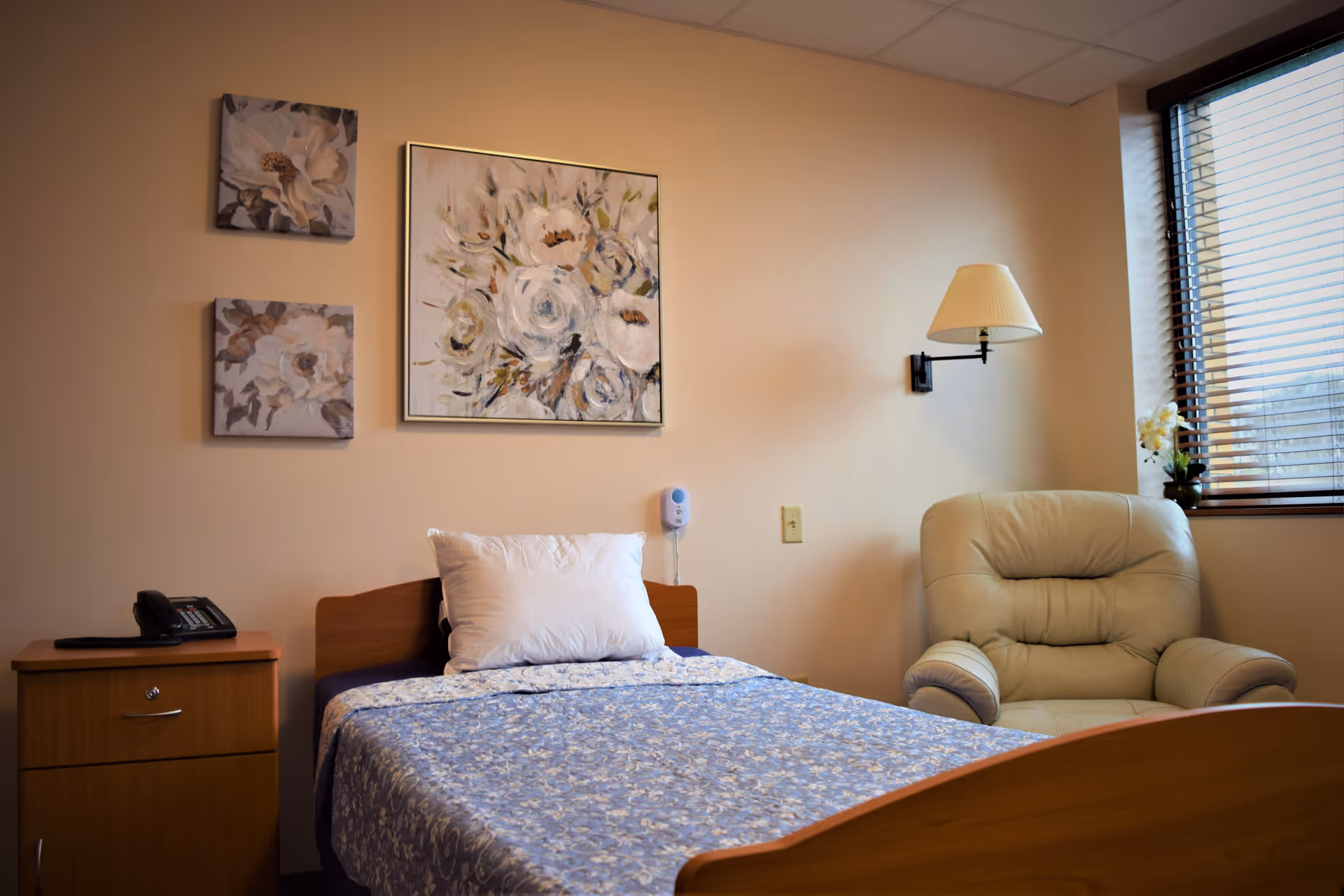 A cozy bedroom in a personal care home featuring a single bed with a blue floral bedspread and a white pillow. Above the bed, there are three floral paintings on the beige wall. To the left of the bed is a wooden nightstand with a telephone on it. To the right, there is a beige cushioned armchair near a window with wooden blinds. A wall-mounted lamp is positioned above the armchair.
