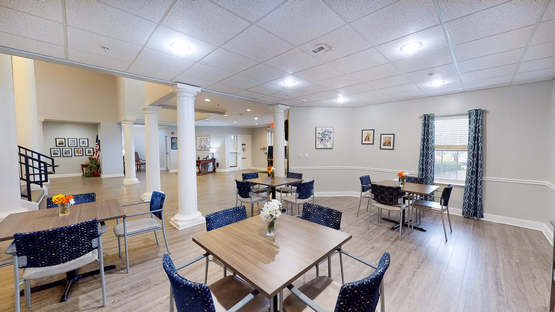 A bright and spacious dining area in a senior living facility with several square wooden tables and blue patterned chairs. The room features light-colored walls, large white columns, wood flooring, framed artwork on the walls, and a window with blue patterned curtains letting in natural light. Small flower arrangements are placed on each table.