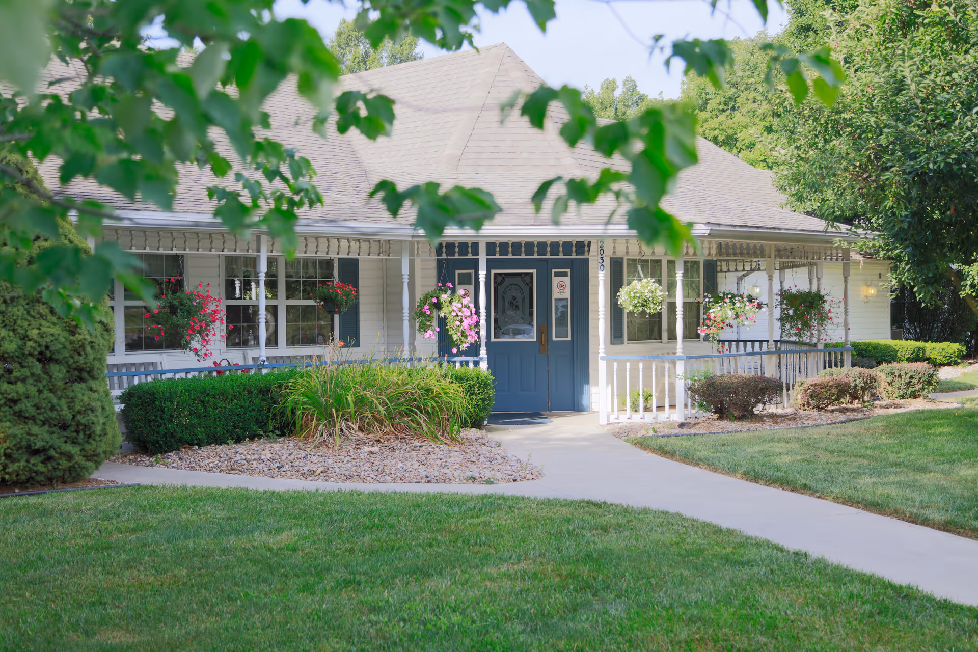 Front exterior view of a single-story building with a blue door, white siding, and a porch decorated with hanging flower baskets and shrubs. A concrete pathway leads to the entrance, surrounded by green grass and trees.