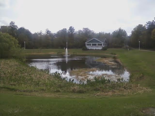 A pond with a central fountain in front of a white porch-front building surrounded by grassy lawns and trees.