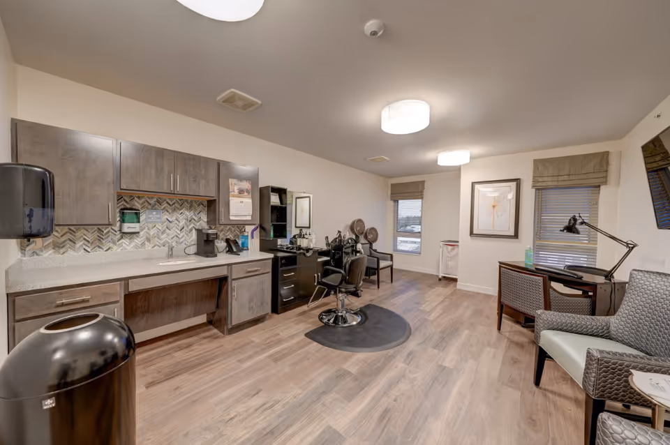 A well-lit salon room in a senior living facility featuring a hair styling chair in front of a mirror with hair care tools, a countertop with cabinets, a coffee maker, and a sink. The room has wood flooring, two windows with blinds, a desk with a lamp, and several chairs for seating.