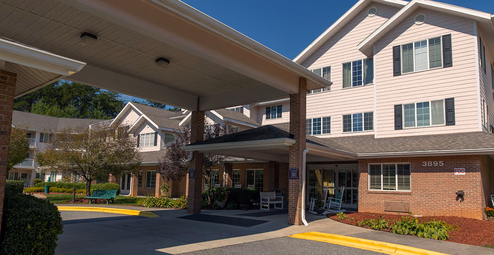 Exterior view of Creekside Terrace senior living facility showing a covered entrance with brick pillars, multiple windows, and a well-maintained garden area with benches and trees under a clear blue sky.