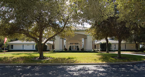 Front exterior view of Brookdale Pointe West facility showing a large entrance with columns, surrounded by trees and a well-maintained lawn under a clear sky.