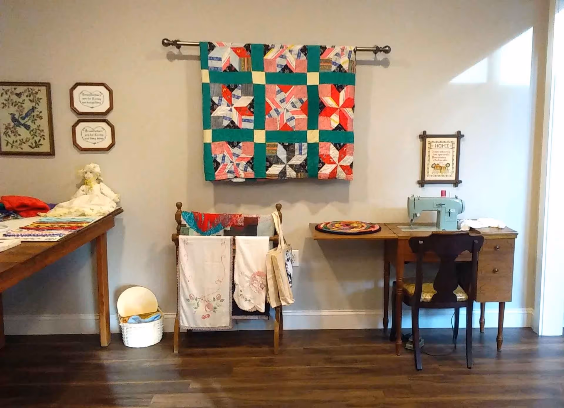 A cozy craft room with a wooden table on the left covered with fabric and craft materials, a quilt rack holding embroidered cloths in the center, and a wooden desk on the right with a vintage sewing machine and a chair. A colorful patchwork quilt hangs on the wall above the quilt rack, and framed decorative signs are mounted on the wall.
