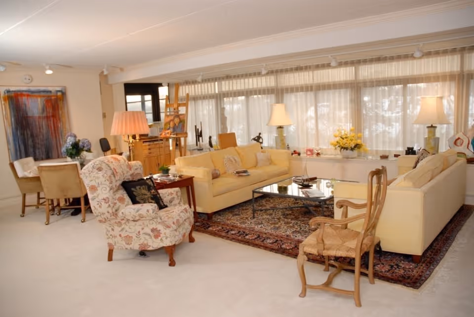 A bright and spacious living room with large windows covered by sheer curtains. The room features two yellow sofas, a floral patterned armchair, a wooden chair, a glass coffee table on a patterned rug, and several table lamps. In the background, there is a small dining area with a table and chairs, a wooden cabinet, and an easel with a painting. The walls are decorated with artwork and flowers.