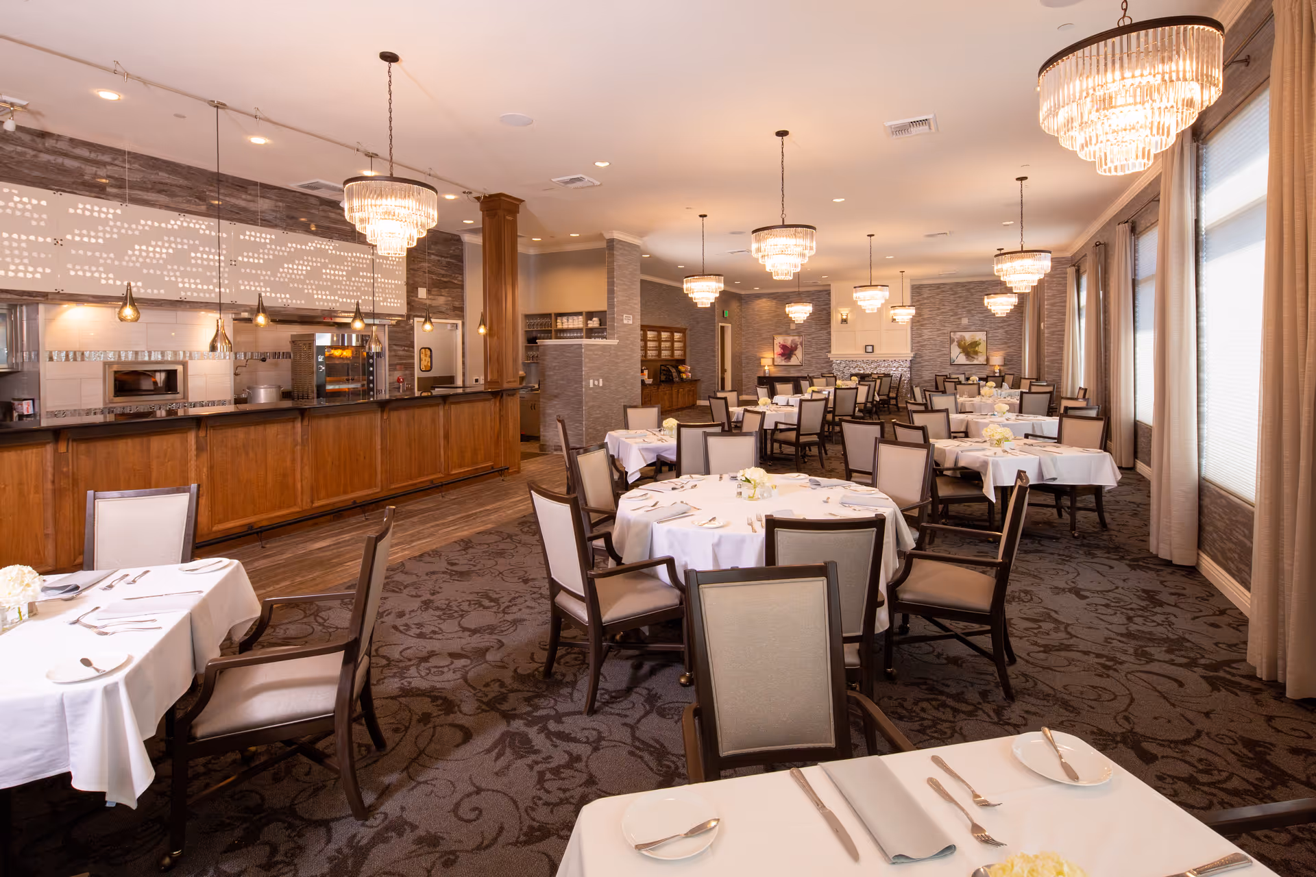 A spacious dining room with multiple round and rectangular tables covered with white tablecloths, set with plates, silverware, and small floral centerpieces. The room features elegant chandeliers hanging from the ceiling, large windows with beige curtains, and a long wooden counter area with pendant lights and kitchen appliances in the background.